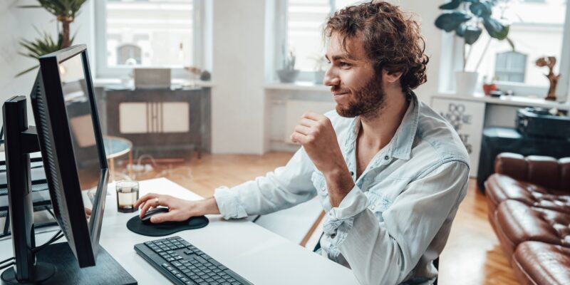 Joyful guy with curly hairs and beard sits at table working on pc